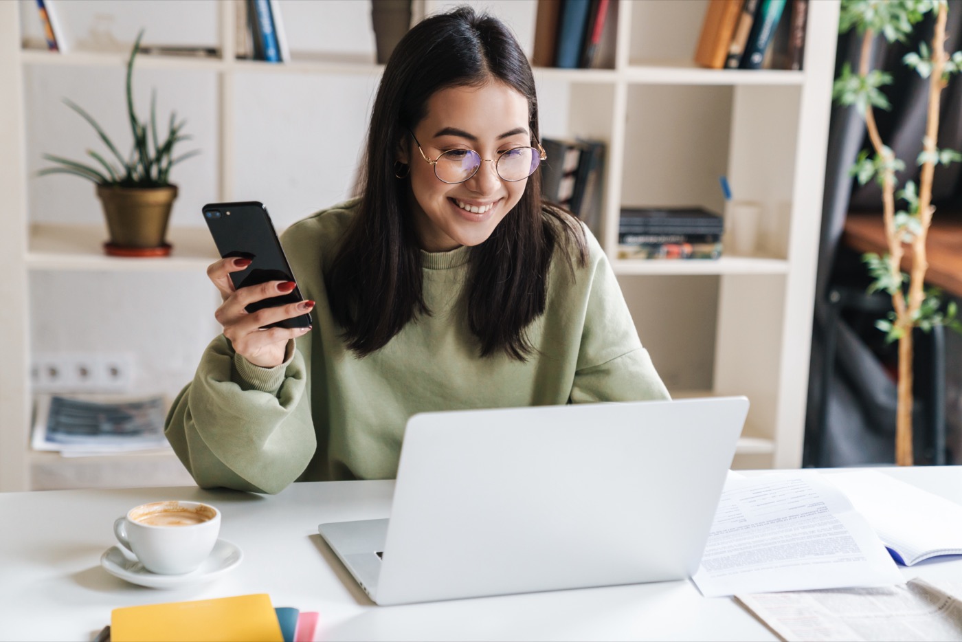 Junge Frau mit Brille hält ein Handy in der Hand während sie am Laptop sitzt